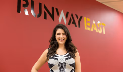 Smiling Natasha Guerra standing in front of Runway East sign on red wall