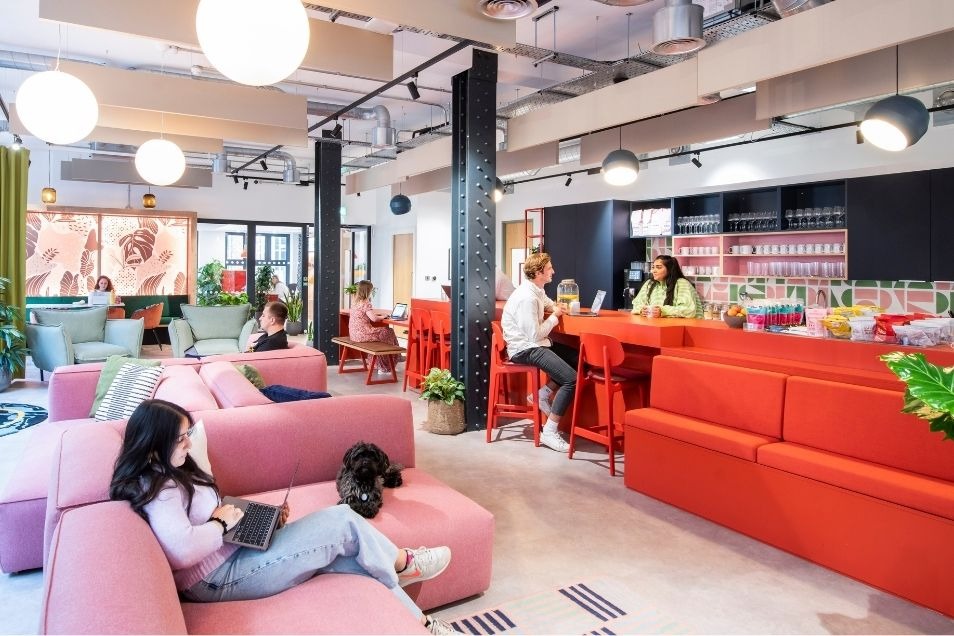 A vibrant, open-plan office featuring pink and red modular seating, an industrial black pillar, and people working in a bright, modern café-style space.