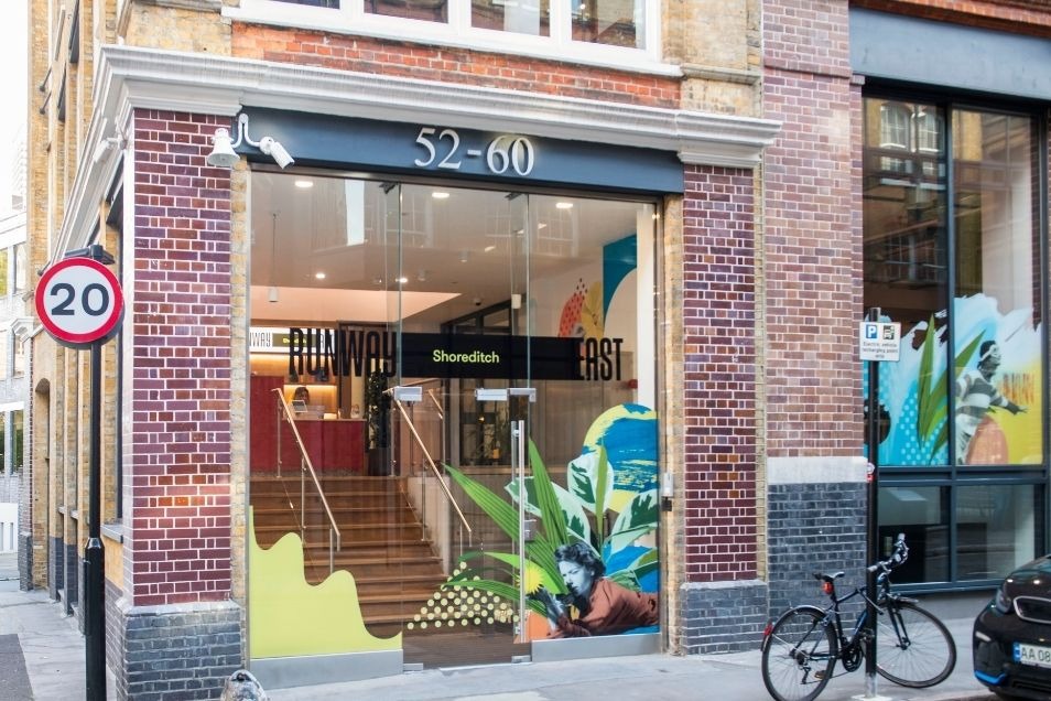 The brick-and-glass exterior of Runway East Shoreditch on a London street corner, featuring colourful window graphics and a "20" speed limit sign.