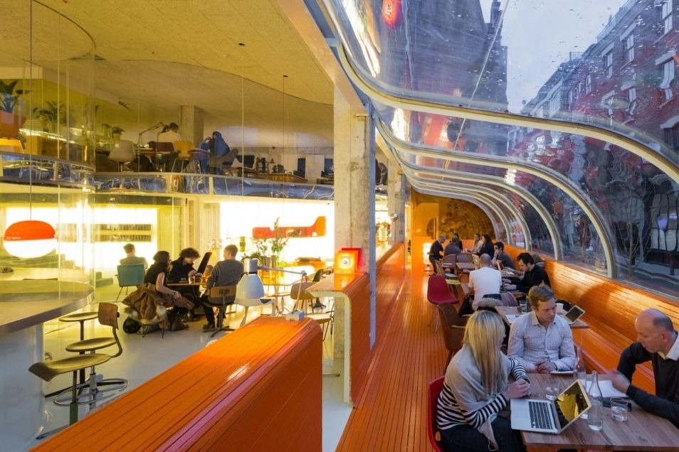 Modern cafe with curved glass walls, orange benches, and people working