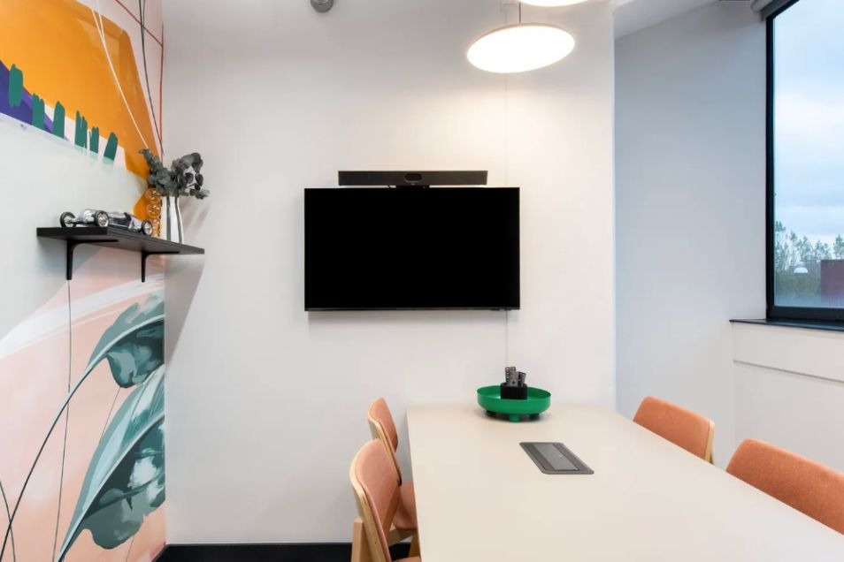 A clean and colourful meeting room with white table, orange chairs, and wall-mounted TV at Runway East Borough Market