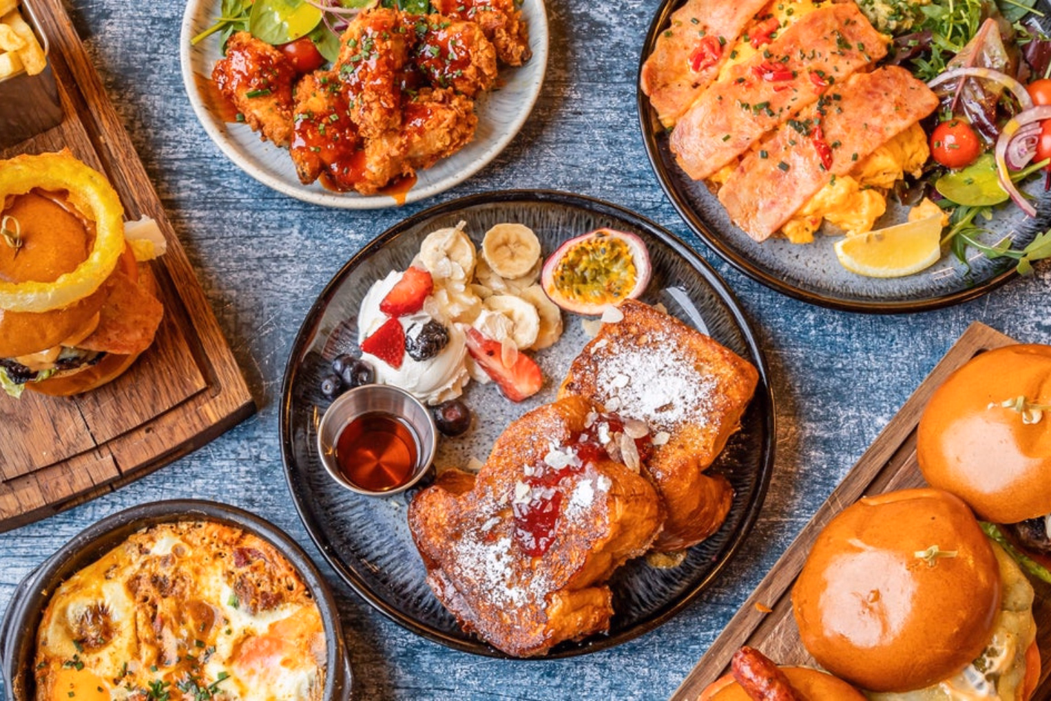 An overhead shot of several brunch dishes including French toast with fruit, pancakes with bacon, fried chicken, and sliders.