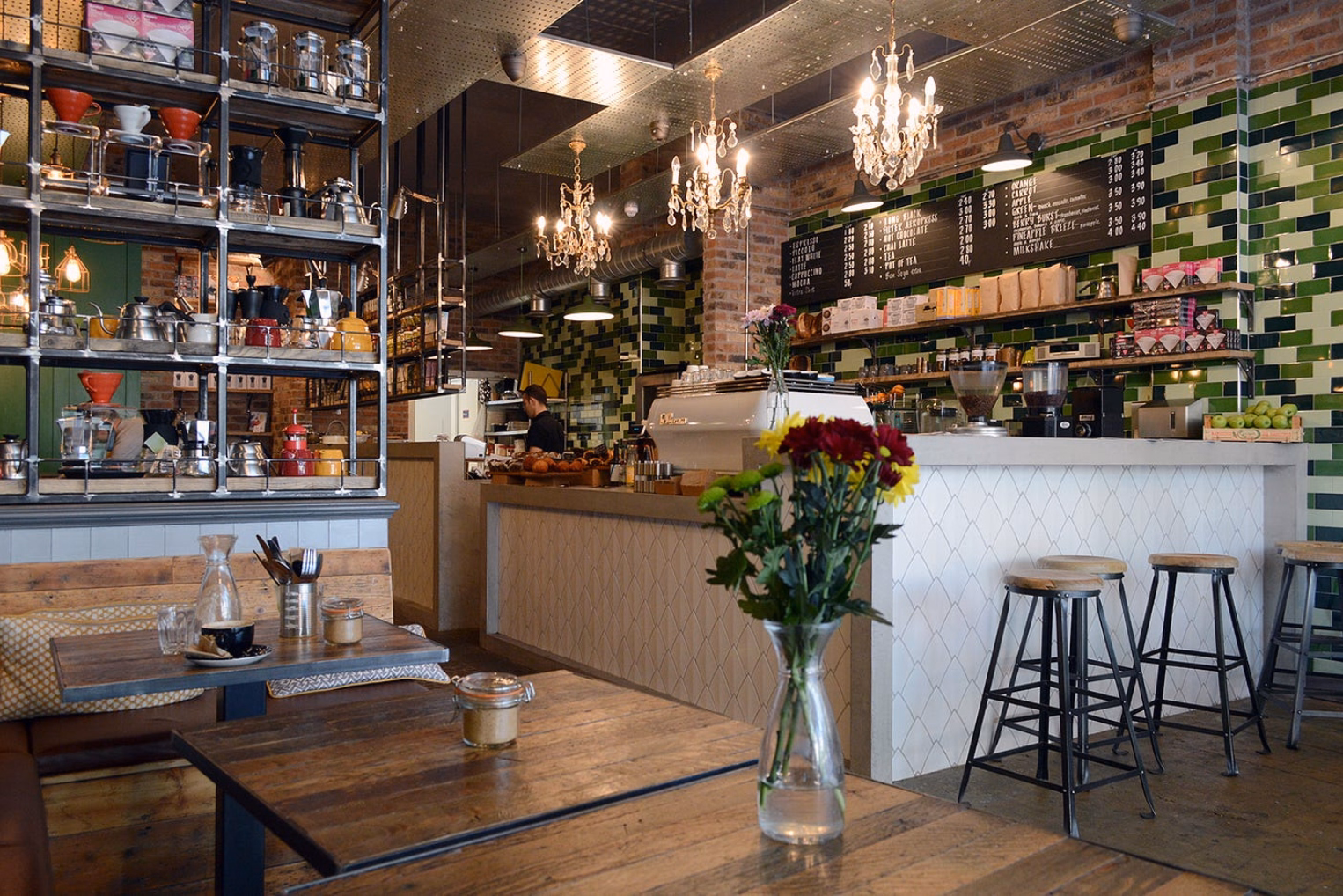 A cozy cafe interior featuring rustic wooden tables, elegant chandeliers, and a white tiled counter with a green tiled backsplash and a chalkboard menu.