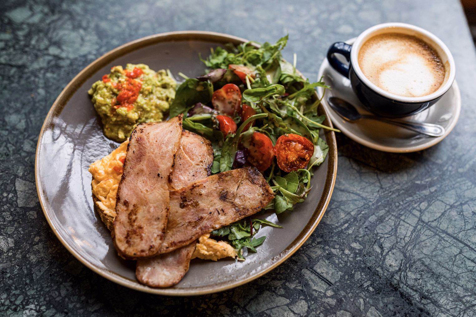 A breakfast plate with thick-cut bacon, scrambled eggs, guacamole, and a side salad, served next to a latte on a green marble table.
