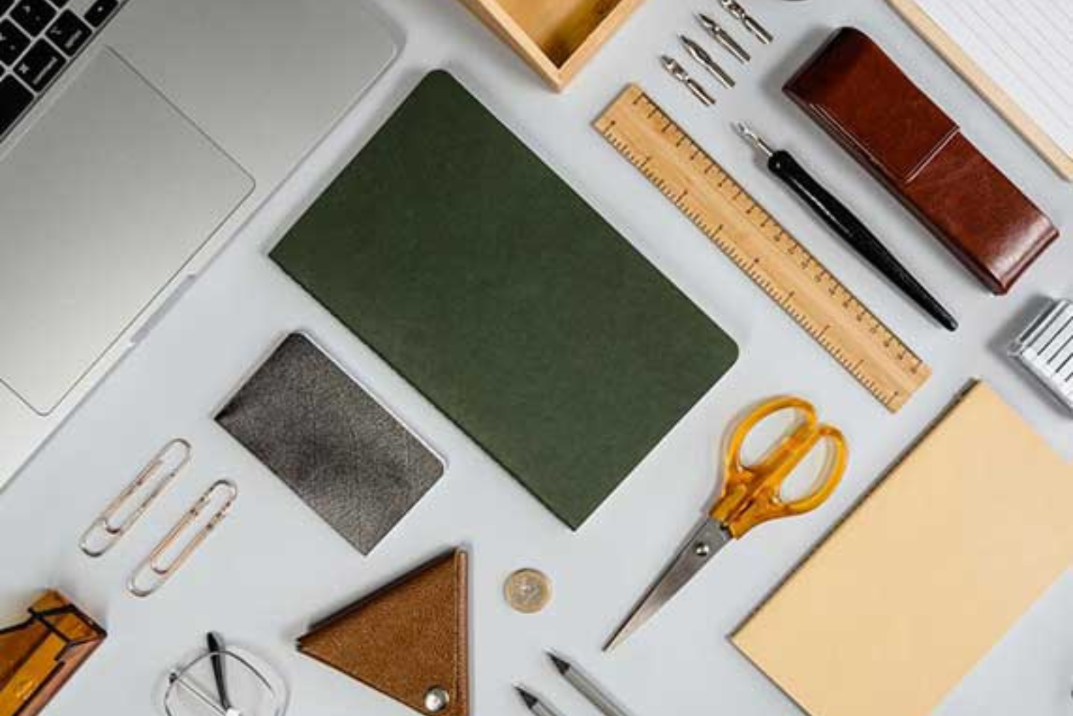 An overhead flat lay of various stationery items, including a green notebook, a wooden ruler, yellow-handled scissors, a leather pencil case, and a laptop corner, all neatly arranged on a white surface.