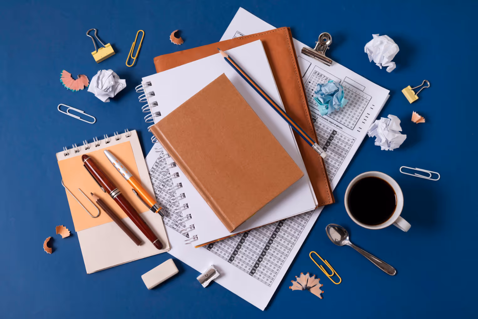 An overhead shot of a messy desk on a blue background, featuring stacked notebooks, loose papers, pens, a pencil, paperclips, and crumpled paper scraps next to a cup of black coffee.
