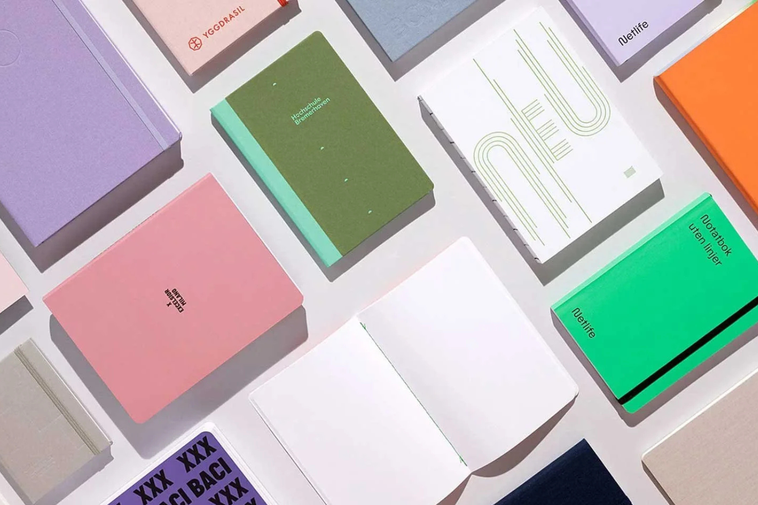 An overhead flat lay of various notebooks in pastel colors, including pink, lavender, green, and orange, arranged diagonally on a light grey surface.