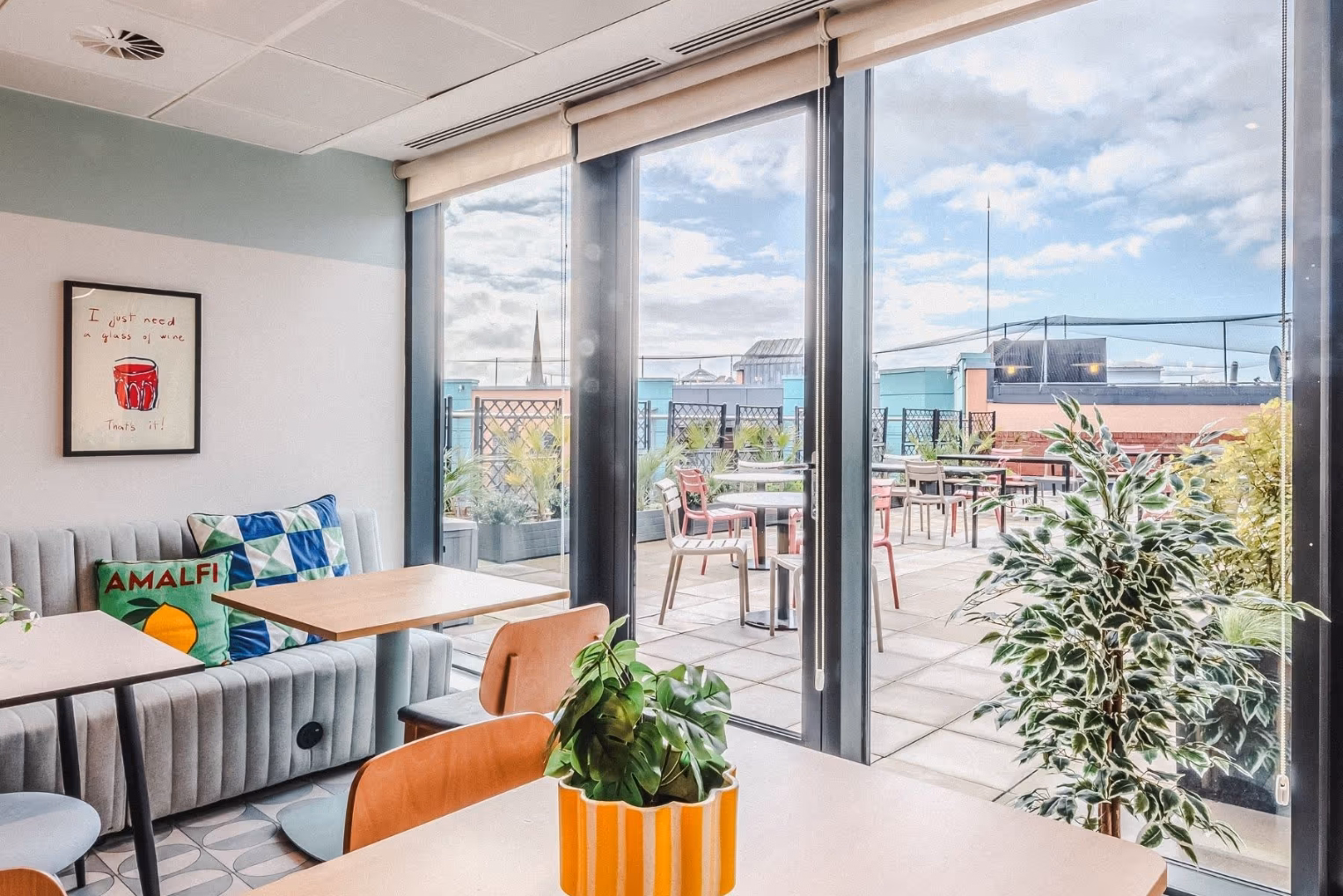 A bright office breakroom featuring a gray fluted banquette with colorful cushions, wood tables, and large glass doors opening onto a sunny rooftop terrace with outdoor seating.