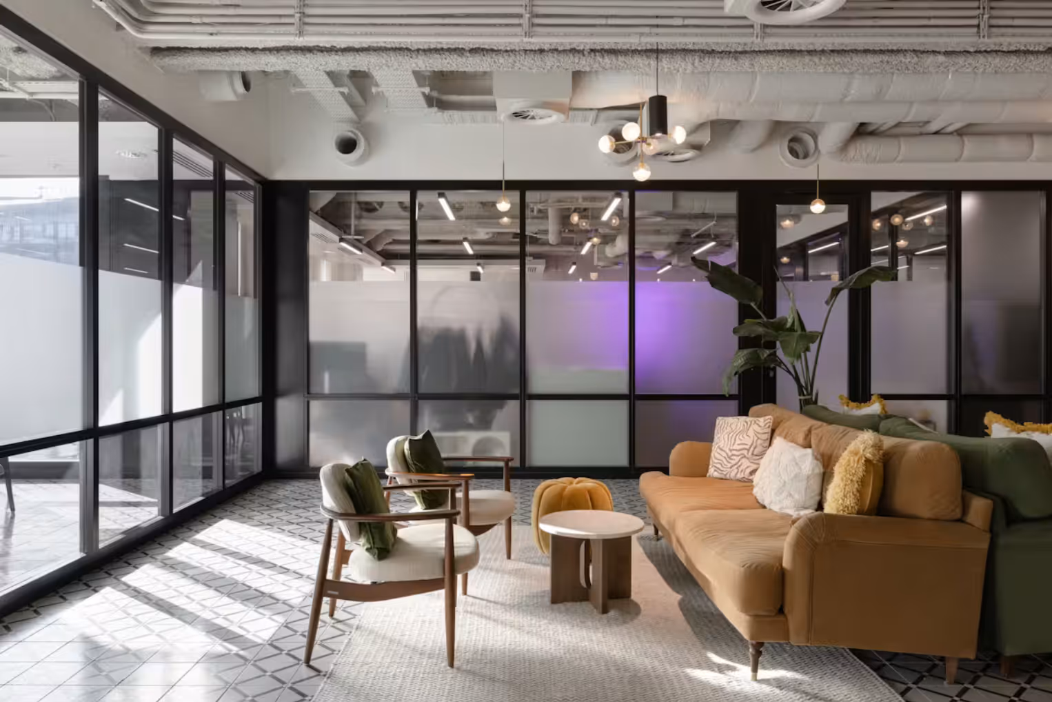 Modern lounge area with a tan velvet sofa, mid-century armchairs, and patterned tile flooring, set against frosted glass partitions and industrial ceilings.