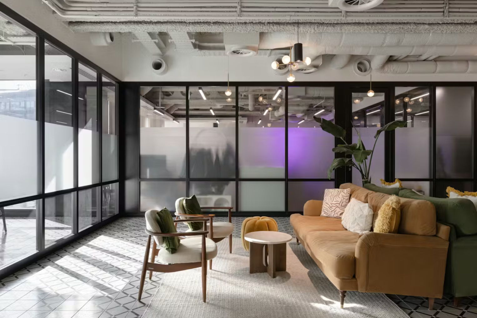 Modern lounge area with a tan velvet sofa, mid-century armchairs, and patterned tile flooring, set against frosted glass partitions and industrial ceilings.