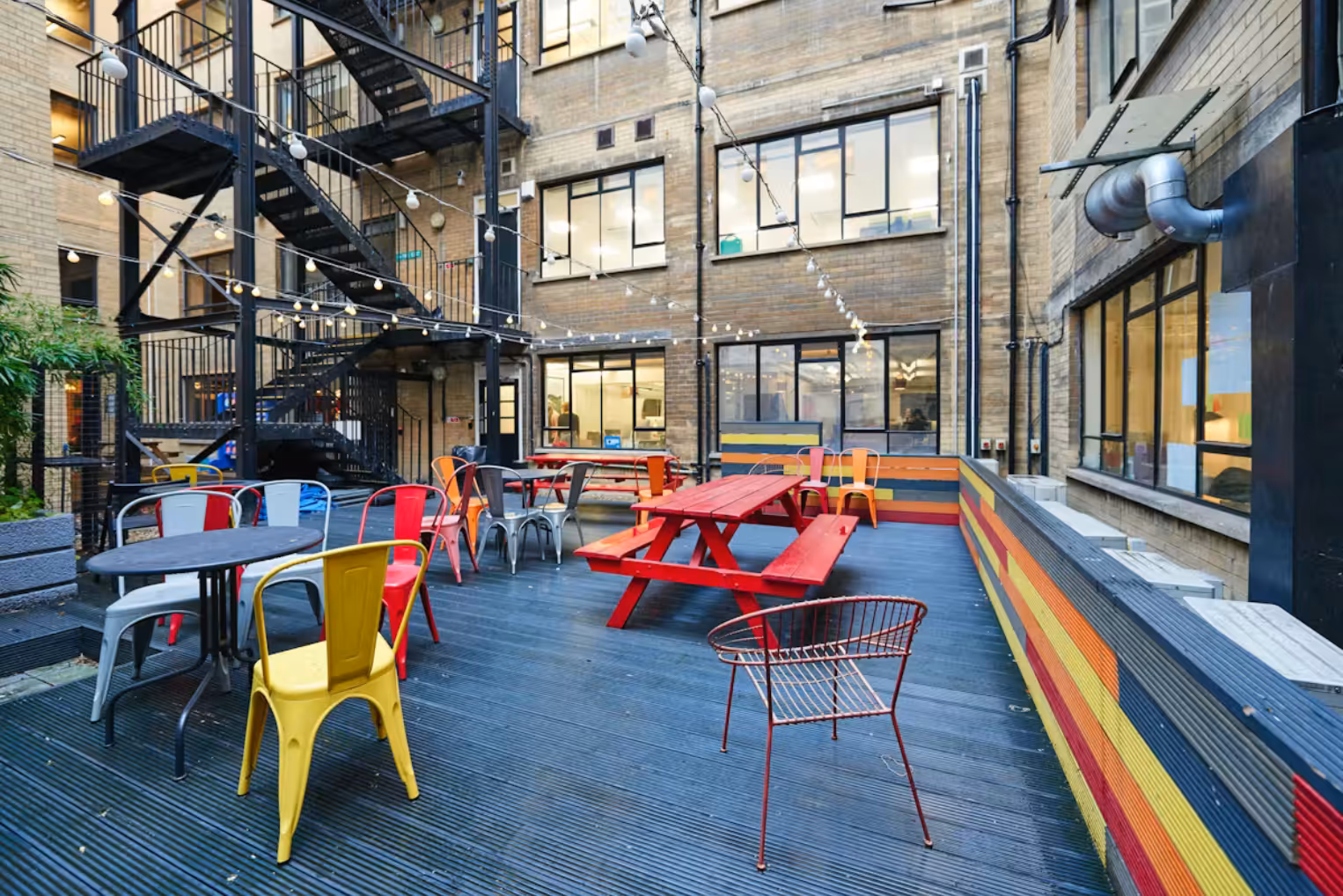A vibrant urban courtyard with red picnic tables, colourful chairs, and string lights set against a brick building with a black metal fire escape.
