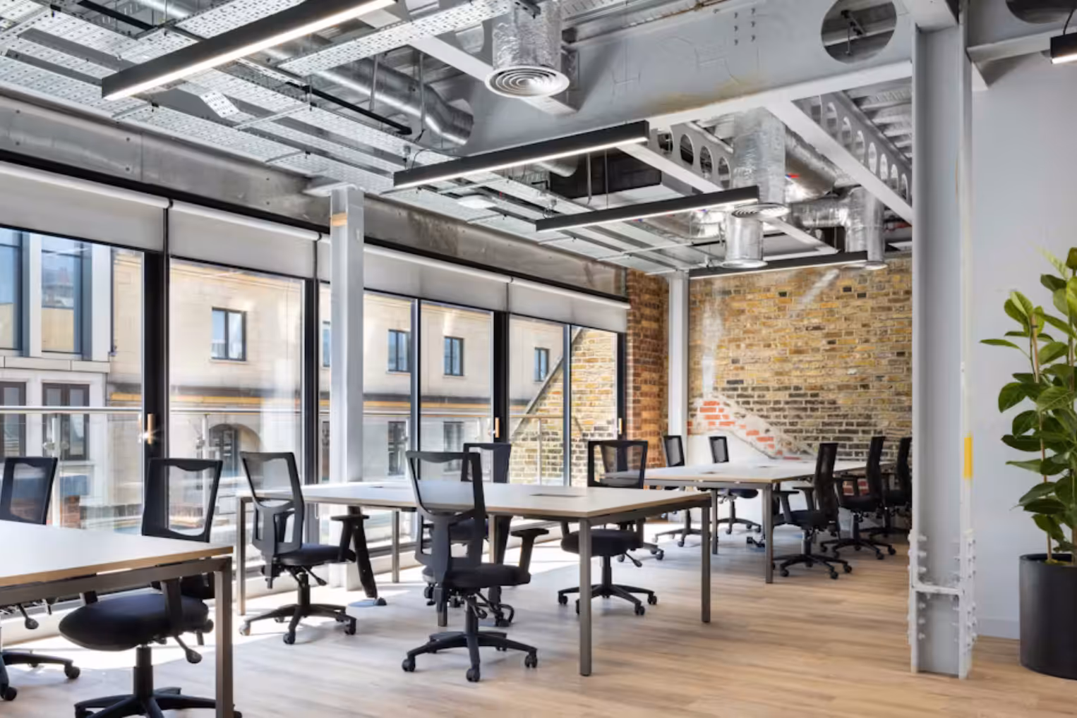 Industrial-style office featuring desks with black ergonomic chairs, large floor-to-ceiling windows, and an exposed brick accent wall.