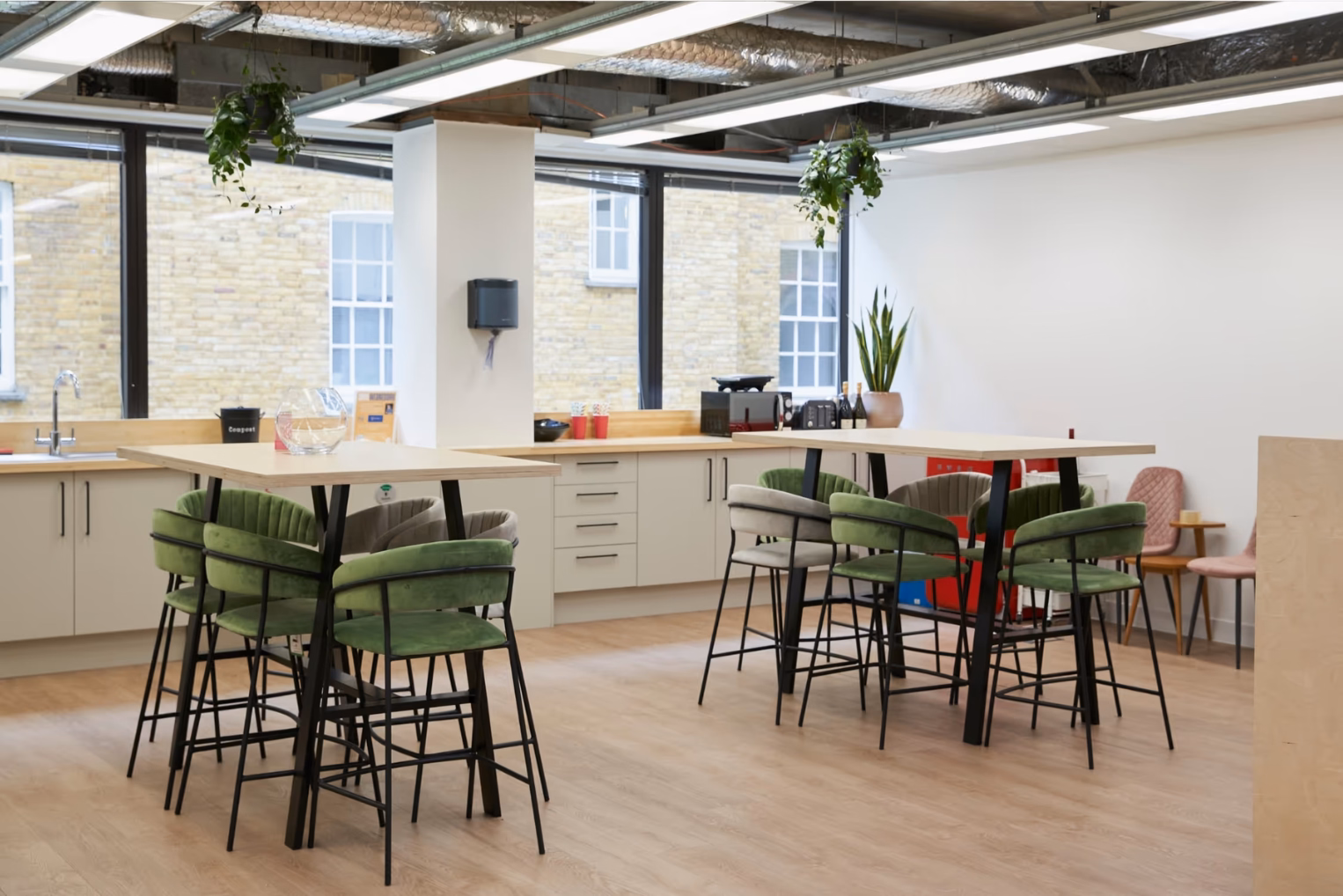 Modern office breakout area featuring high wooden tables with green velvet stools, a minimalist kitchenette, and large windows.