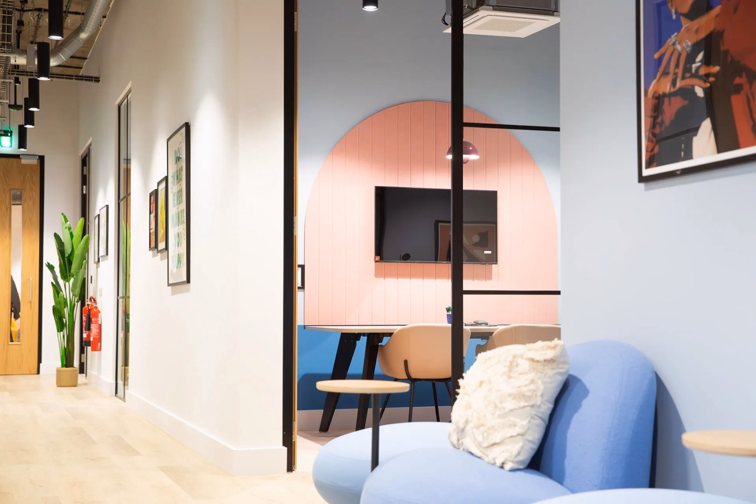 Office corridor leading to a meeting room with a pink arched wall panel, blue seating, and minimalist wall art.
