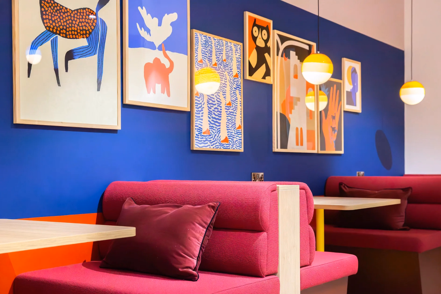Vibrant office booth seating with bright pink sofas against a royal blue wall adorned with a gallery of colourful, abstract framed prints.