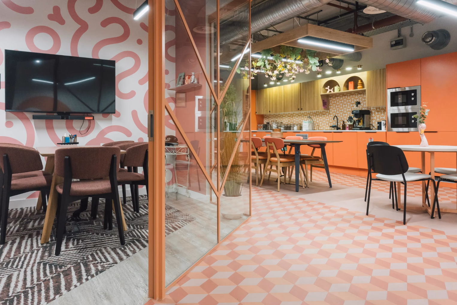 A playful office breakout area and kitchen featuring pink squiggly wall art, geometric floor tiles, and orange cabinetry with industrial exposed ducting.