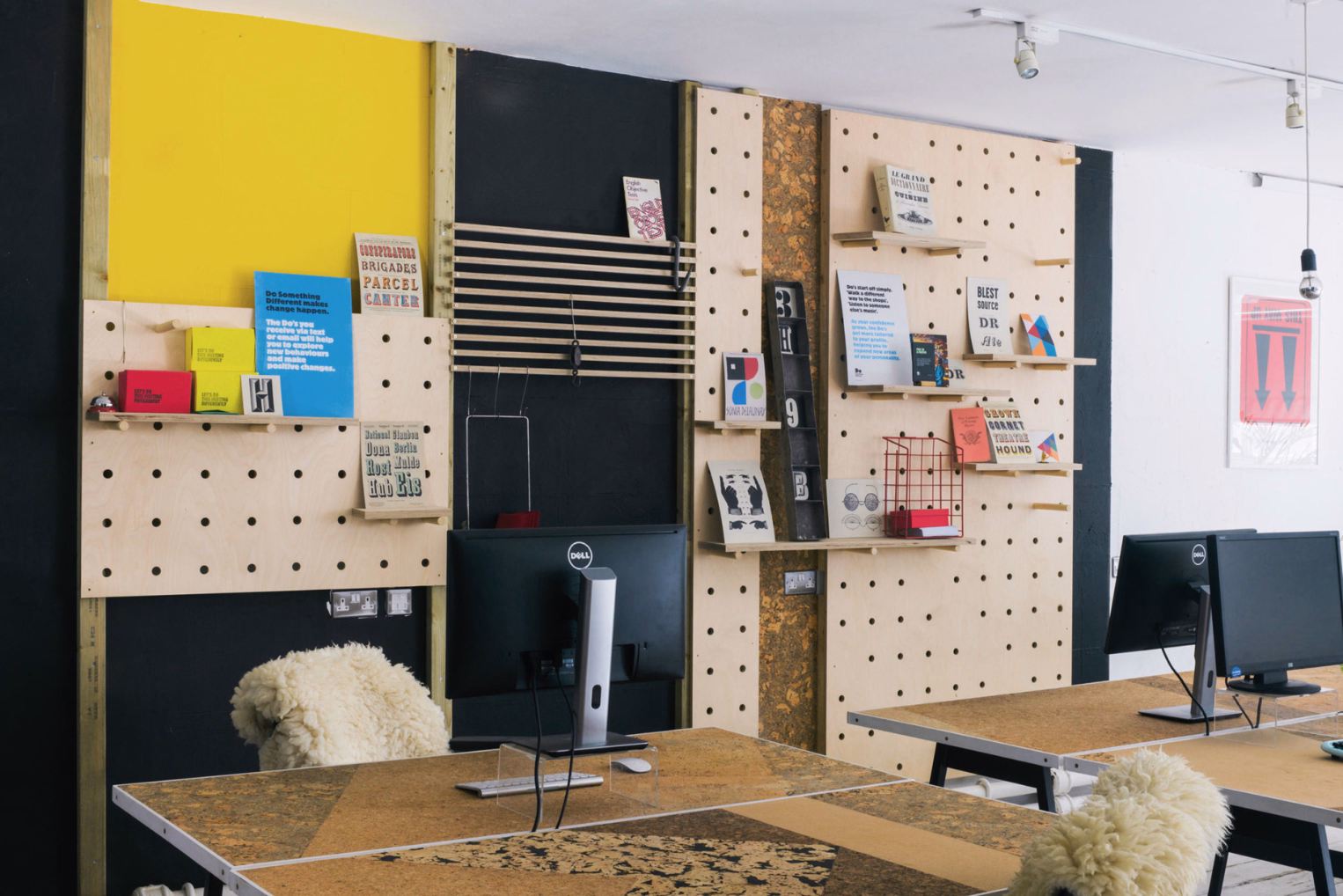 Creative office workspace featuring cork-top desks, sheepskin chair covers, and large plywood pegboards decorated with various prints and books.