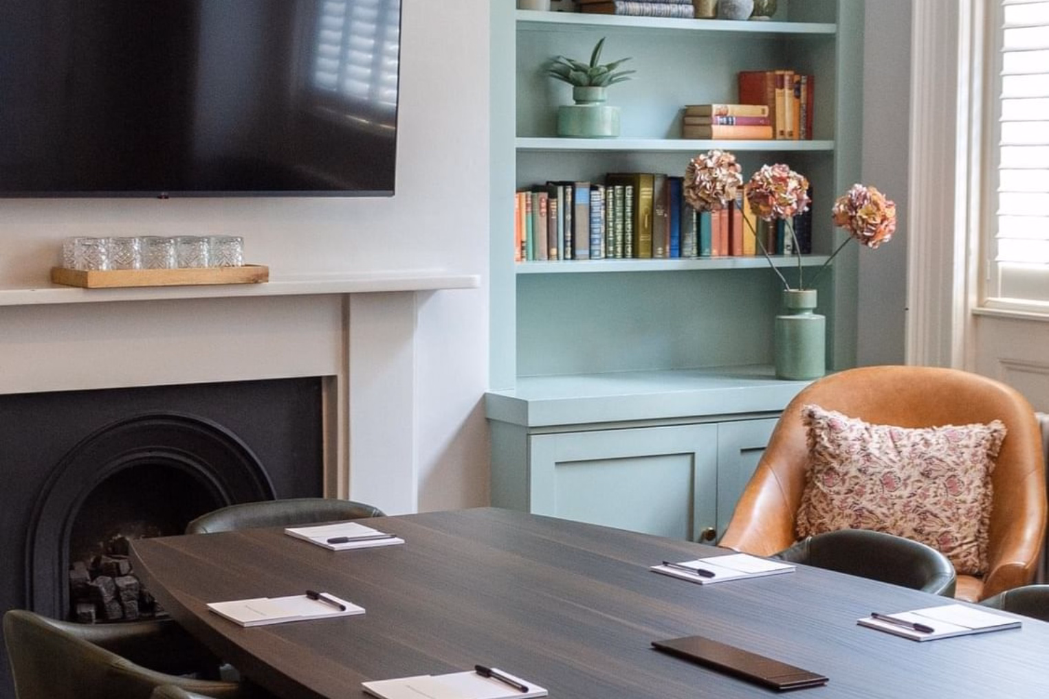 Elegant meeting room featuring a dark wood table with note pads, a classic fireplace, and a sage green built-in bookshelf filled with books.