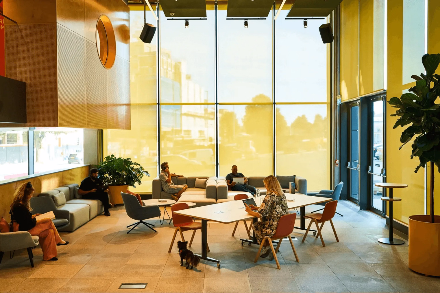 Bright, high-ceilinged office lobby with yellow-tinted windows, featuring people working on modular sofas and a large white table.