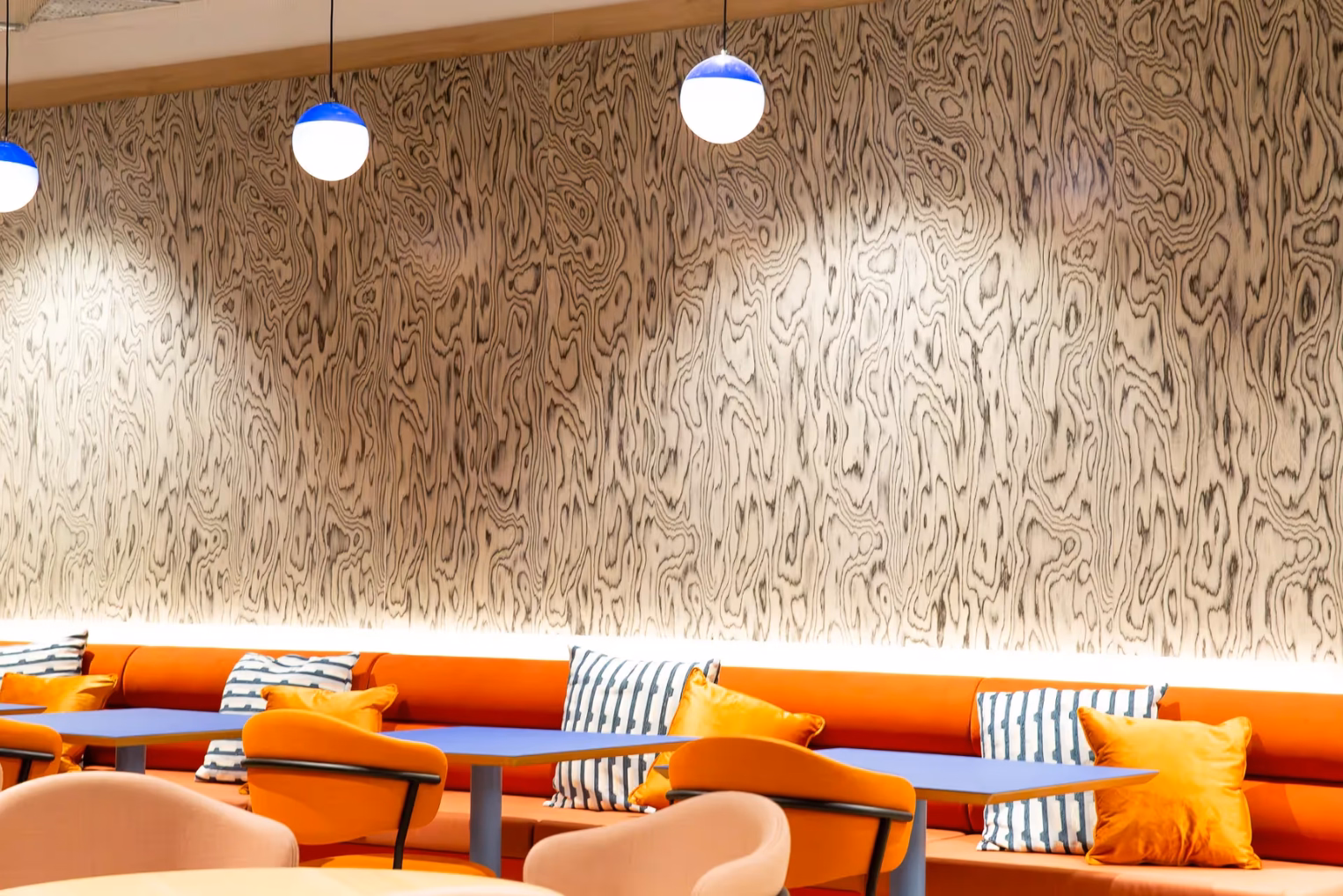Seating with bright orange banquettes, blue tables, and spherical pendant lights against a striking wood-grain patterned wall.