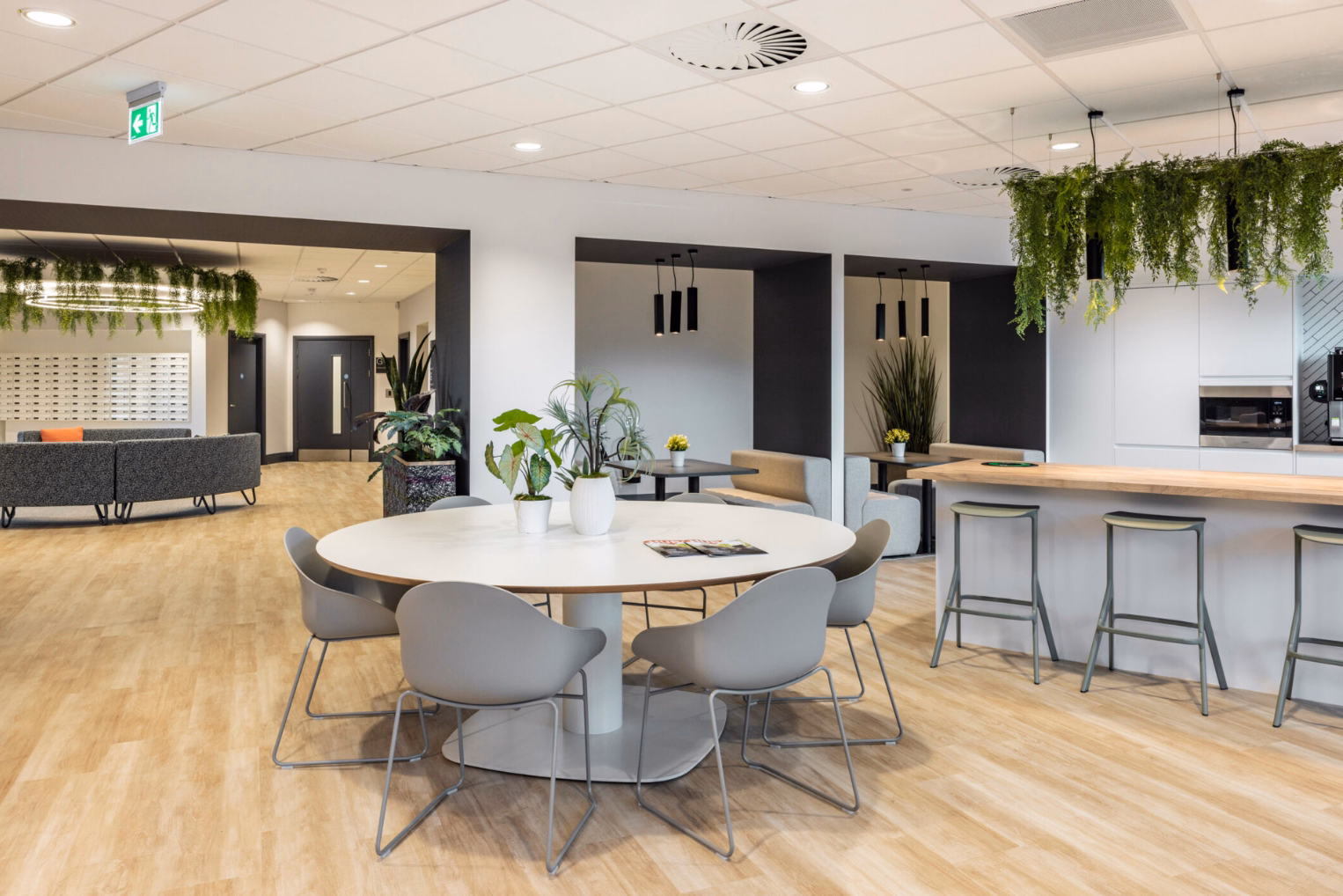 Office breakroom featuring a white round table, kitchen island with bar stools, and grey booth seating recessed into black-lined alcoves.