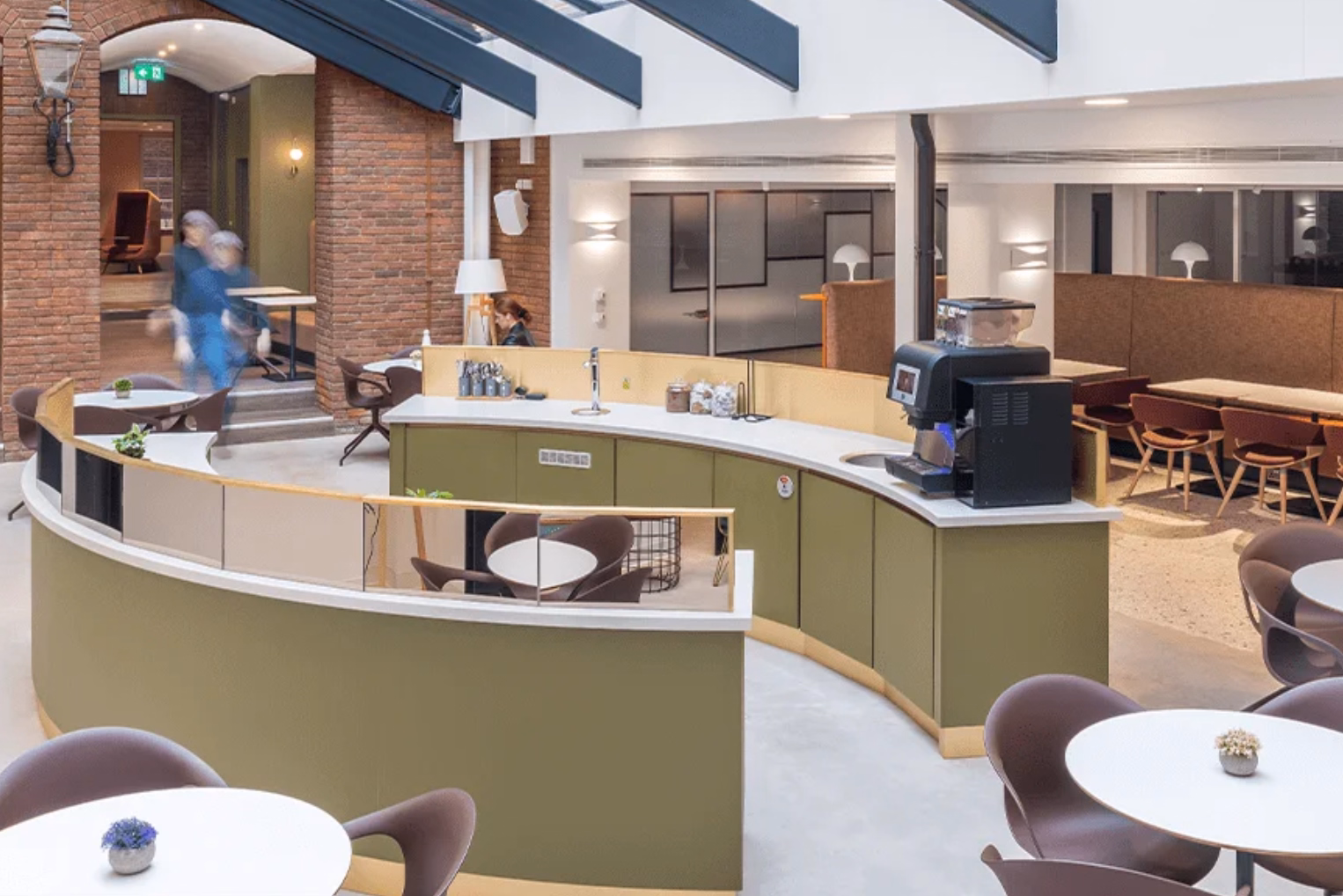 Modern office atrium with a curved olive green coffee bar, white cafe tables, and exposed brick walls beneath a skylight ceiling.