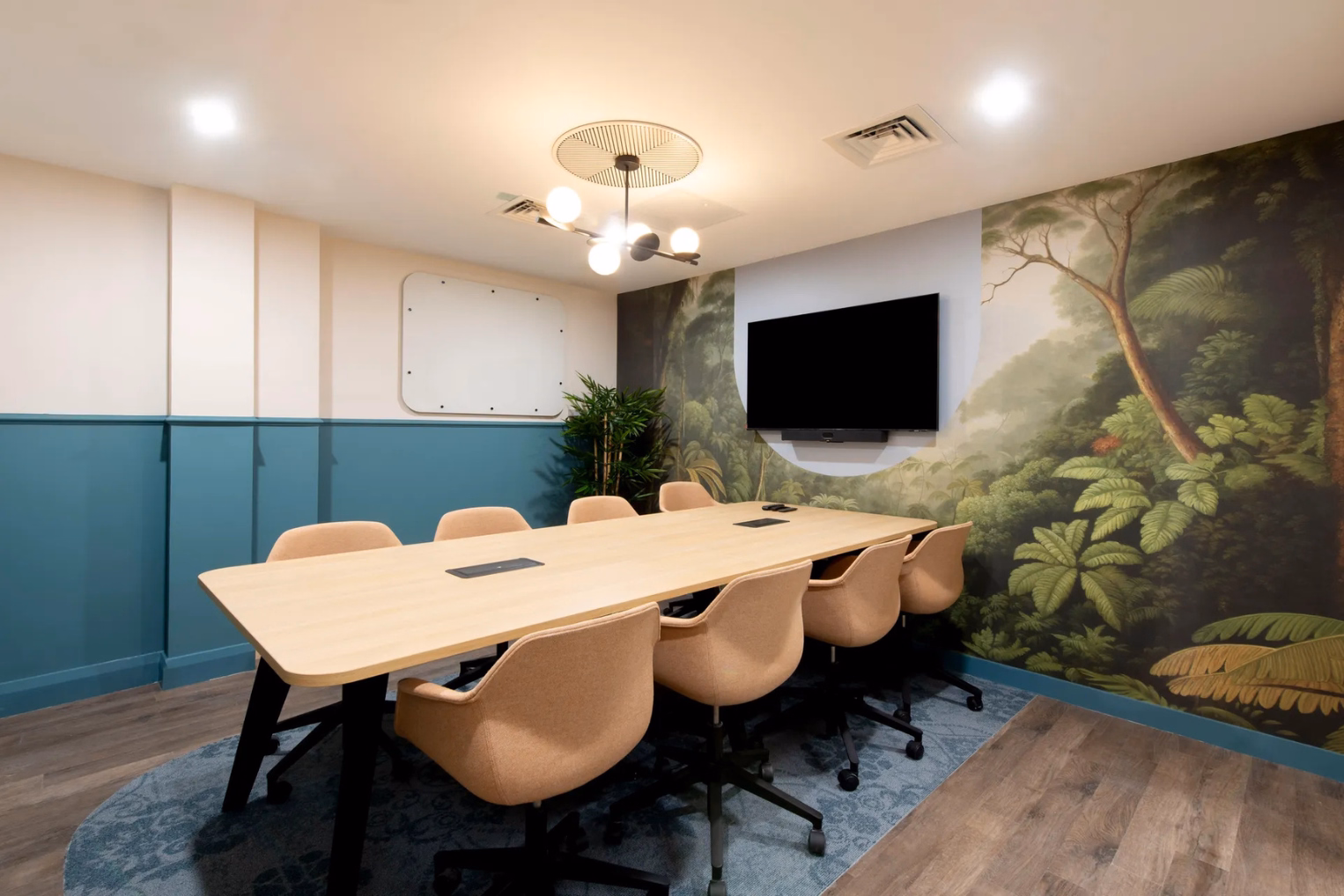 Modern meeting room featuring a wooden table with tan chairs, a teal wainscoted wall, and a large tropical jungle mural.