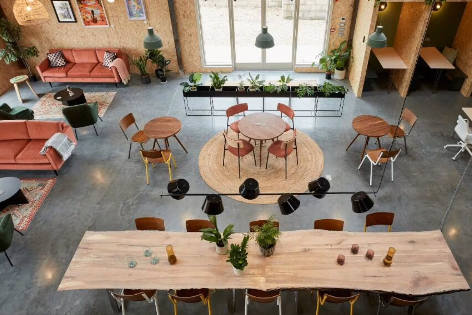 High-angle view of an industrial workspace with coral sofas, a live-edge wooden table, and private plywood booths on a polished concrete floor.