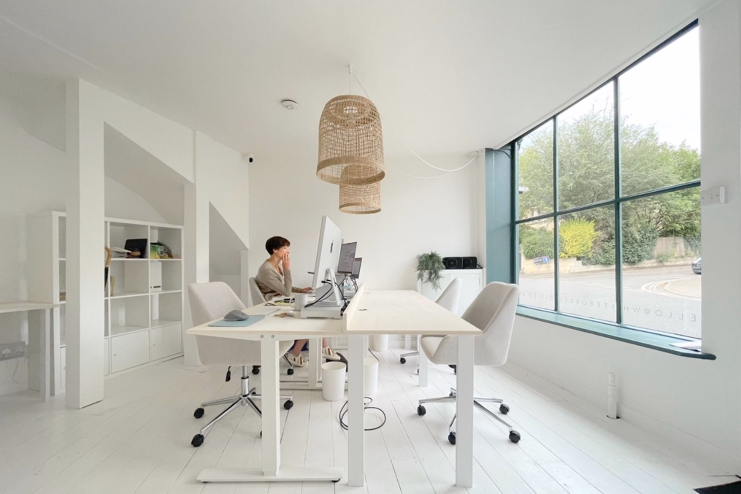Minimalist all-white office featuring a person at a long desk, rattan pendant lights, and large windows overlooking a green street.