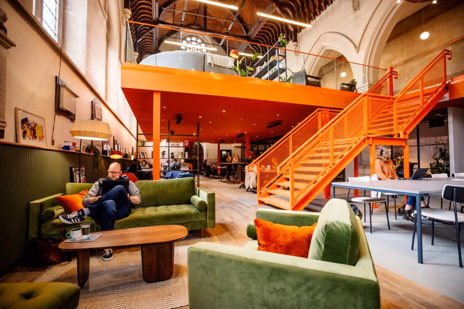 A vibrant coworking space in a converted church with a bold orange mezzanine, green velvet sofas, and original vaulted ceilings.