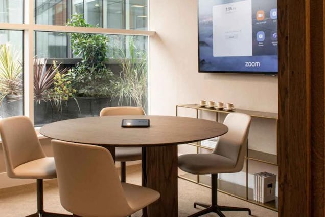 Small circular meeting room with a wooden table, tan chairs, a Zoom display, and a window overlooking greenery.