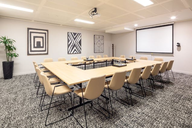 Spacious conference room with a U-shaped wooden table, tan chairs, geometric wall art, and a large projection screen.