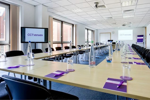 Large conference room set up in a U-shape with light wood tables, black chairs, water bottles, and screens displaying "CCT Venues."