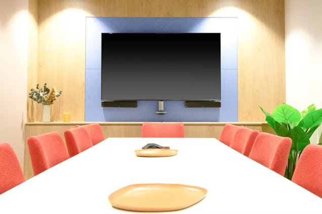Meeting room with a white table, coral-coloured chairs, and a large TV screen set against a blue and wood-panelled wall.