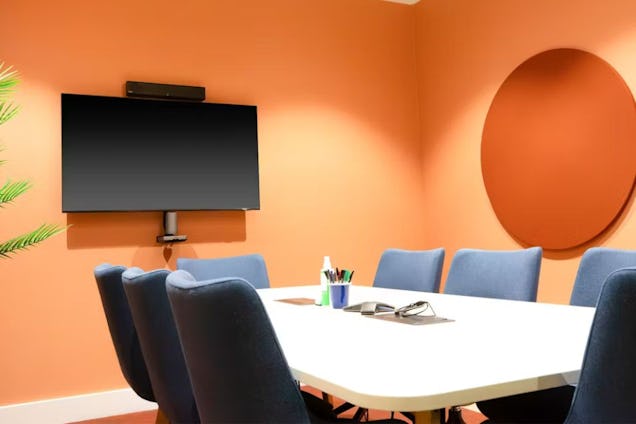 Meeting room with a white table, navy blue chairs, orange walls, and a large wall-mounted monitor.