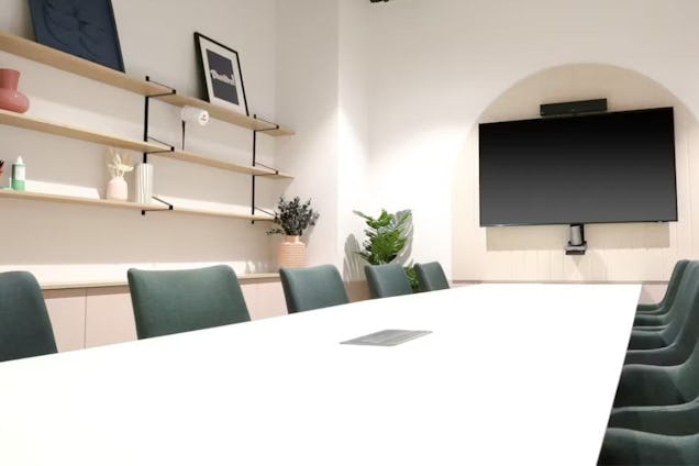 Bright meeting room with a white table, dark green chairs, open shelving with decor, and a TV mounted in a recessed arch.