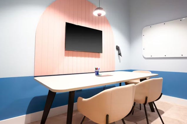 Meeting room with a white table, cream chairs, blue and white walls, and a TV mounted on a pink arched slatted panel.