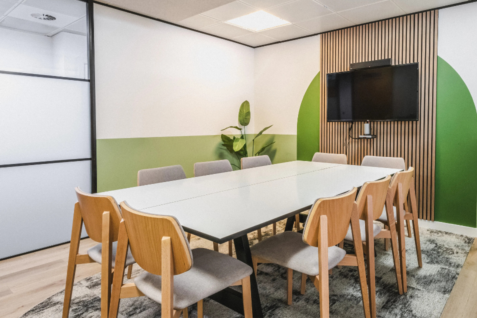 Meeting room with a white table, wooden chairs, a green-and-white wall, and a TV on a slatted wood panel.