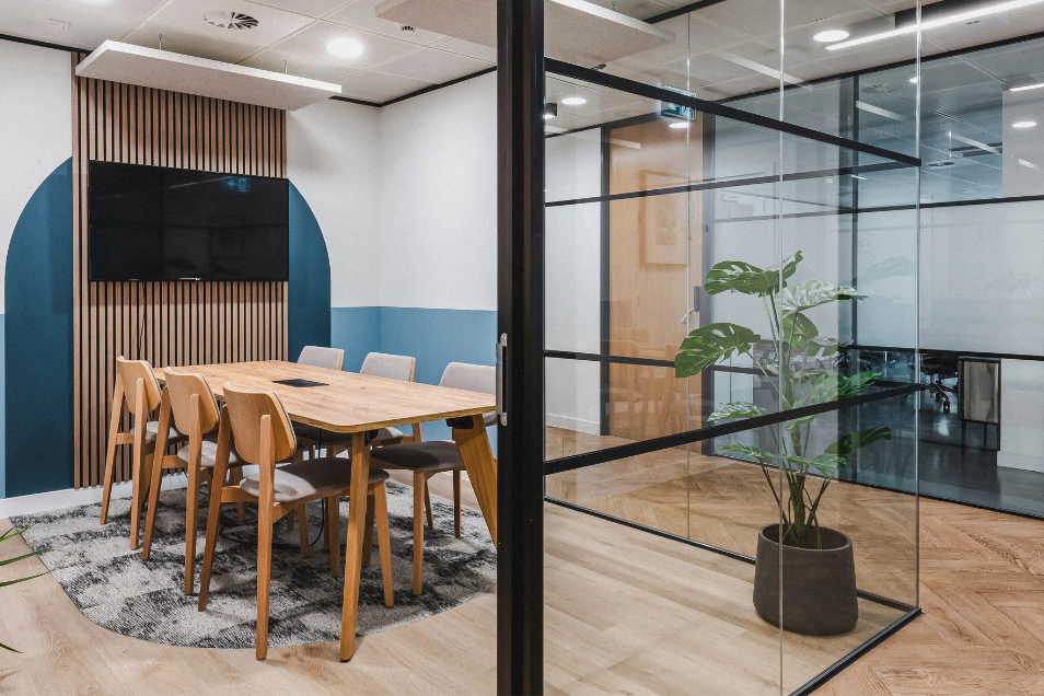 Meeting room with wood furniture and blue walls, viewed through a black-framed glass partition beside a large indoor plant.