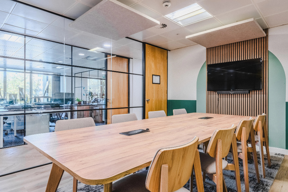 Meeting room with a light wood table, slatted wall panel, and a black-framed glass partition looking into a bright office.