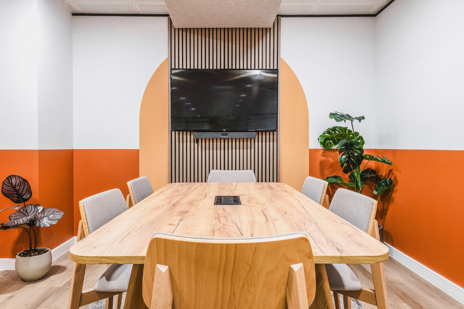 Meeting room with a light wood table, orange and white walls, indoor plants, and a TV mounted on a slatted wood panel.