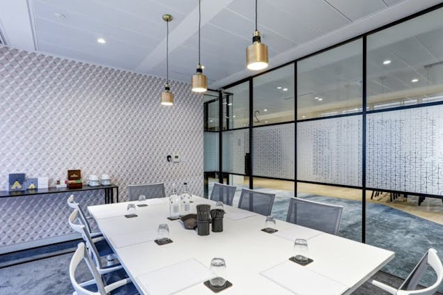 Meeting room with a white rectangular table, mesh chairs, brass pendant lights, and a black-framed glass partition wall.