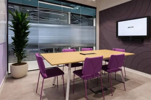 Meeting room with a light wood table, vibrant purple chairs, a tall indoor plant, and a glass-walled partition.
