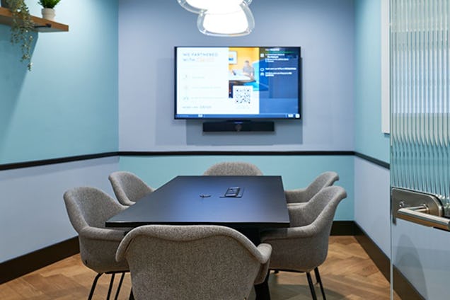 Compact meeting room with a black table, grey bucket chairs, blue-toned walls, and a wall-mounted screen.