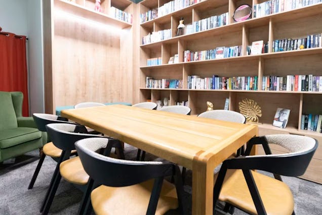 Cosy library-style meeting room with a wooden table, black-framed chairs, and extensive floor-to-ceiling bookshelves.