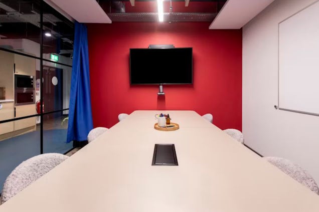 Meeting room with a long white table, a bold red accent wall, a wall-mounted display, and a vibrant blue privacy curtain.