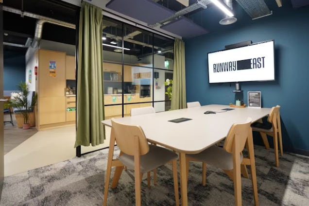 Meeting room with light wood chairs and table, a deep blue accent wall, and a glass partition with olive green curtains.