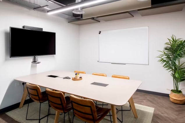 Meeting room with a light wood table, tan leather-backed chairs, a large whiteboard, and a wall-mounted display.