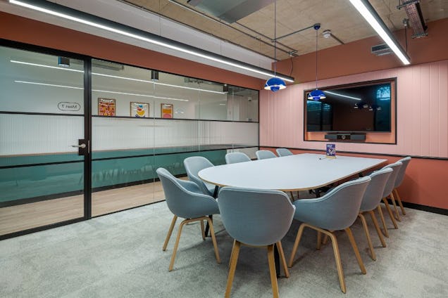 Meeting room with light blue bucket chairs, an oval white table, dusty rose walls, and a glass-panelled partition wall.