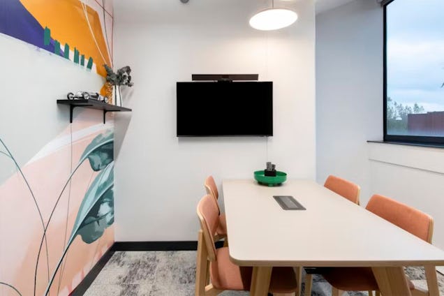 Meeting room with peach-coloured chairs, a light wood table, a wall-mounted screen, and a vibrant botanical mural.